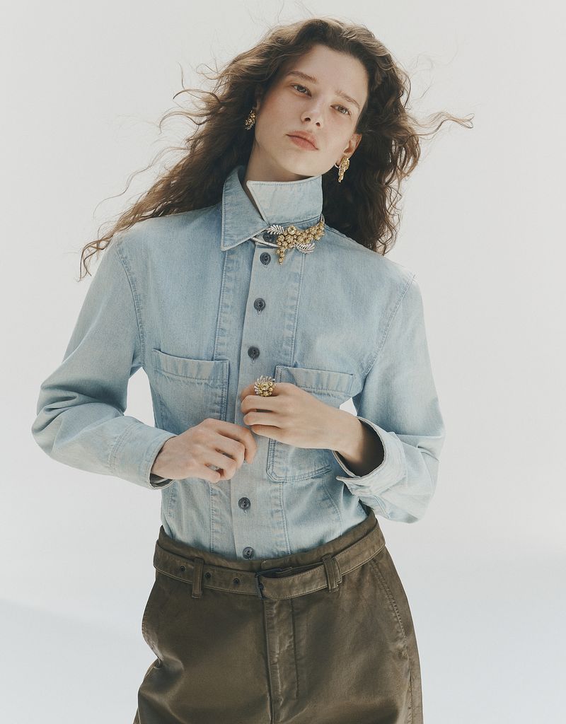 Model wearing a light blue denim shirt and brown trousers with a high-end jewelled collar brooch against a plain white background