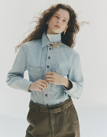 Model wearing a light blue denim shirt and brown trousers with a high-end jewelled collar brooch against a plain white background