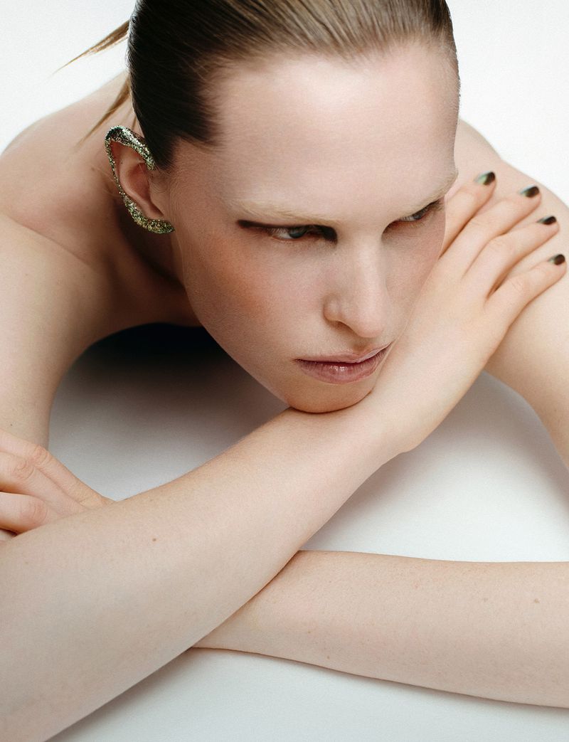 Close-up portrait of a model resting chin on crossed arms, wearing a textured ear cuff against a clean white background.