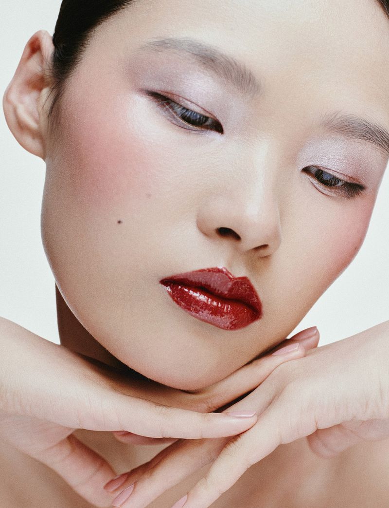 Close-up beauty portrait of a model with glossy burgundy lips, rosy blush, and soft eyeshadow against a white background