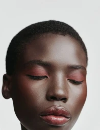 Close-up beauty portrait of a Black woman with short hair and closed eyes wearing soft red eyeshadow and glossy lips against a white background.