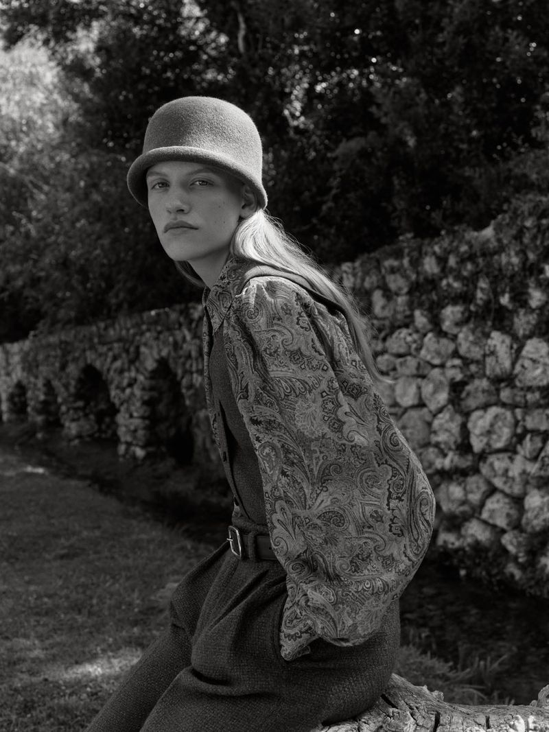 Monochrome fashion portrait of a model wearing a felt cloche hat and patterned blouse against a stone wall