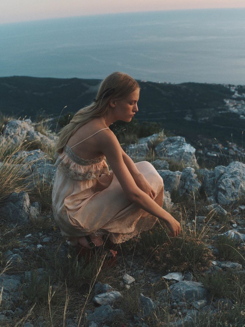 Woman in pale pink dress crouching on a rocky mountain slope overlooking the sea for Manifesto Magazine