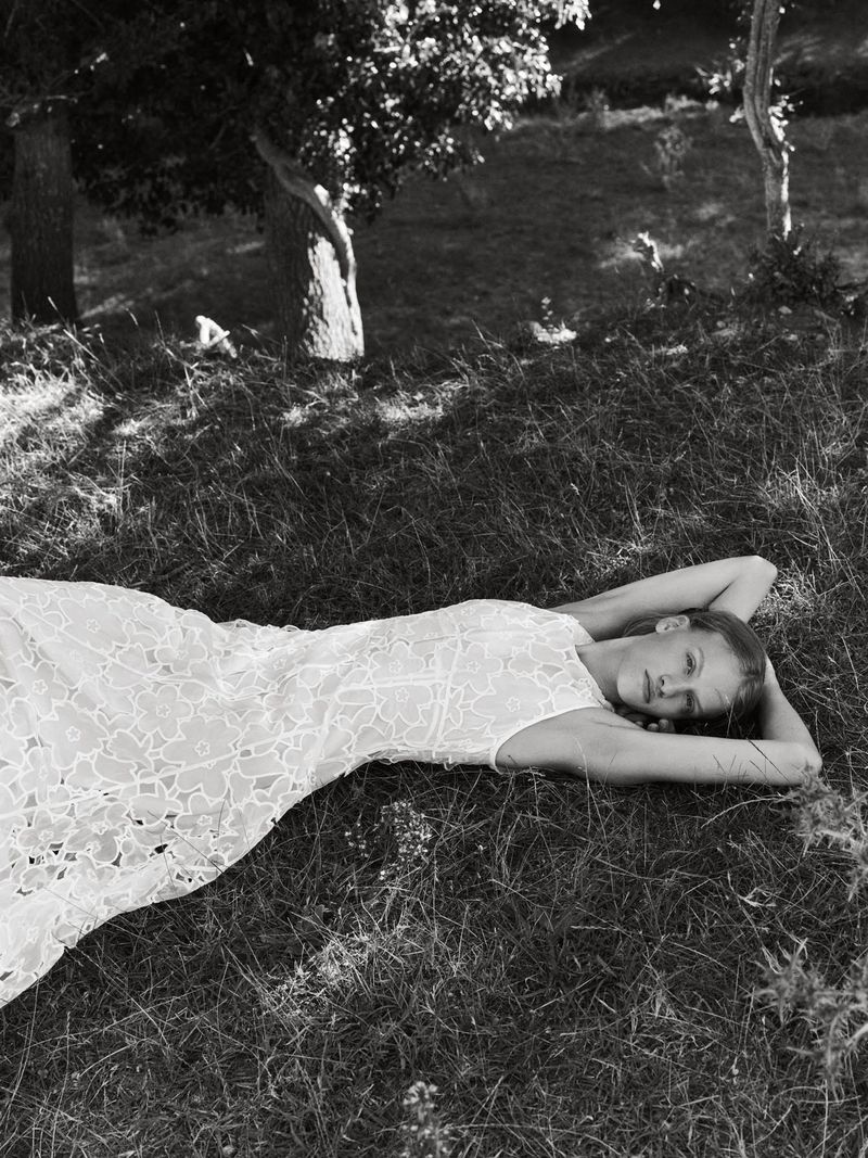 Model lying down in a grassy field wearing a patterned lace dress with arms raised behind her head in black and white