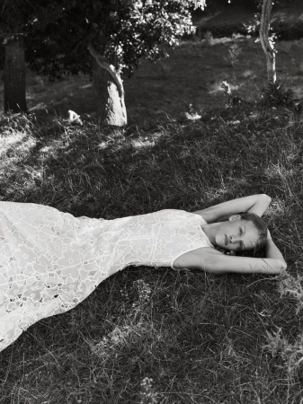 Model lying down in a grassy field wearing a patterned lace dress with arms raised behind her head in black and white