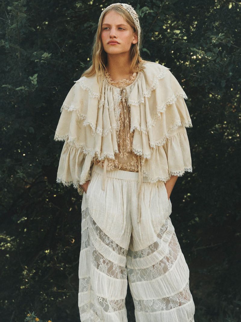 Model in layered cream lace cape and matching trousers standing outdoors before dark foliage in a portrait by Sam Wilson.