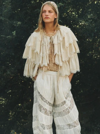 Model in layered cream lace cape and matching trousers standing outdoors before dark foliage in a portrait by Sam Wilson.