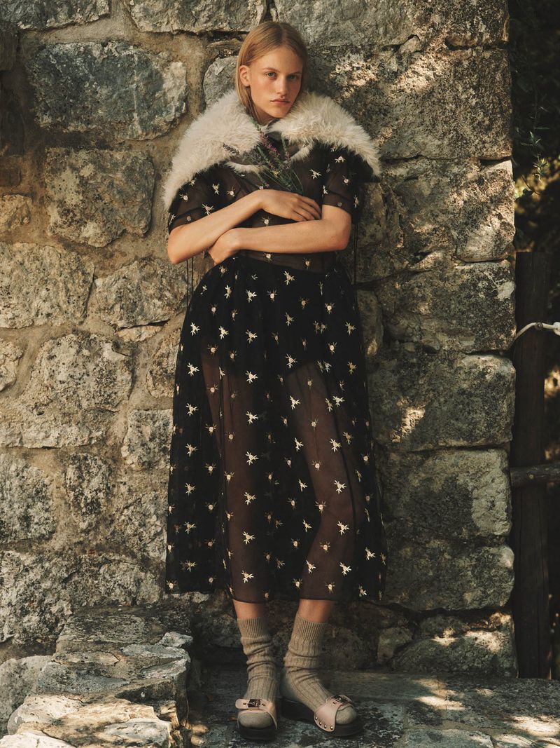 Model leaning against a rustic stone wall wearing a patterned black dress, fur stole, socks, and platform shoes.