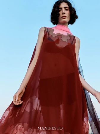 Model wearing a sheer red dress and pink scarf standing against a bright blue sky for Manifesto Magazine
