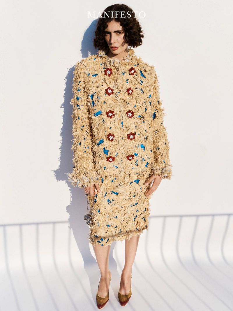 Model wearing a textured cream fringed coat with brown floral buttons standing against a white studio wall with shadows.