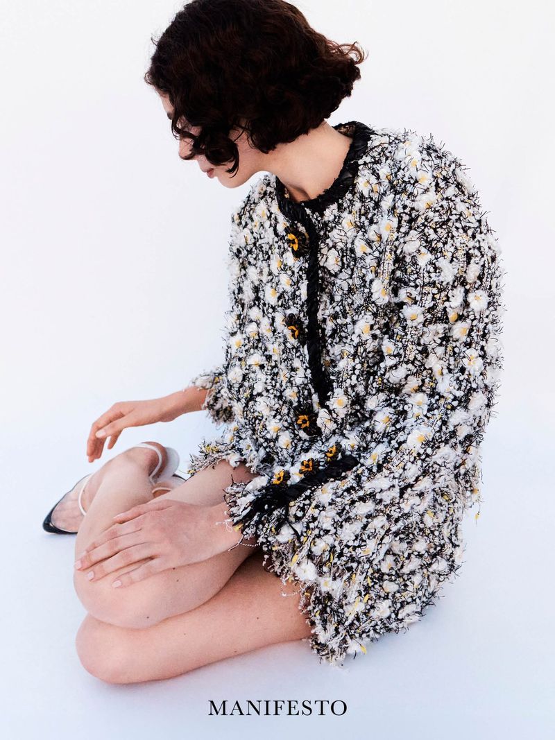 Model with dark curly hair wearing a textured tweed jacket seated against a plain white background