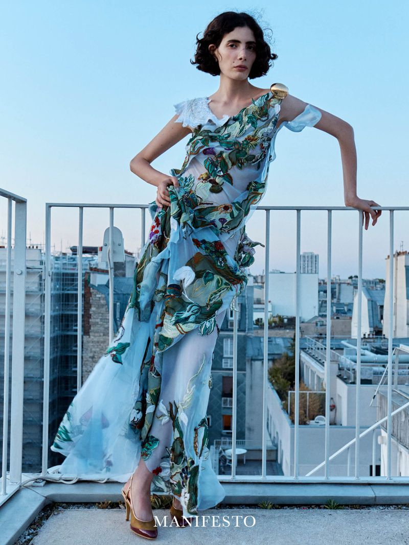 Model in a floral printed blue dress leaning against a rooftop railing with city buildings in the background.