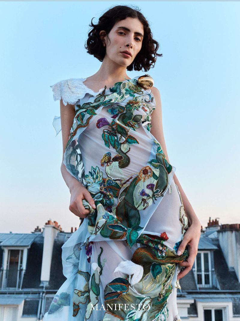 Model wearing an embroidered sheer dress against a rooftop and blue sky background for Manifesto Magazine editorial.