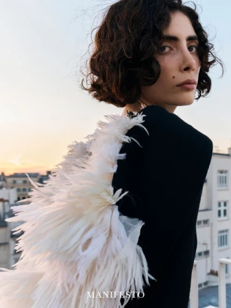 Woman in black top with white feather accessory looking over her shoulder against an urban rooftop at sunset