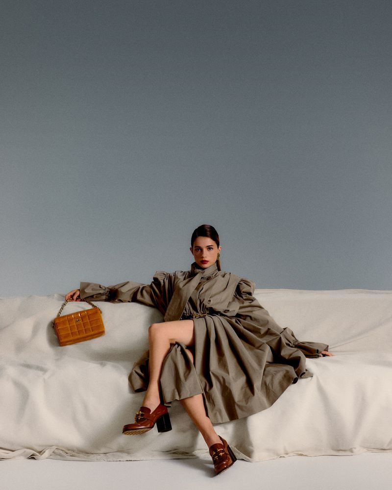 Actress Lisa-Marie Koroll seated on a draped white sofa wearing a grey trench coat, brown loafers, and holding a yellow quilted handbag.