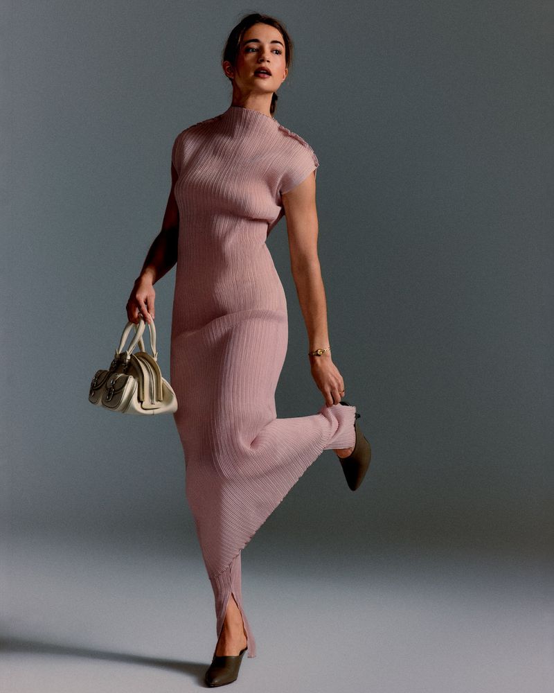 Full-length portrait of actress Lisa-Marie Koroll wearing a textured pink sleeveless dress and holding a white handbag.