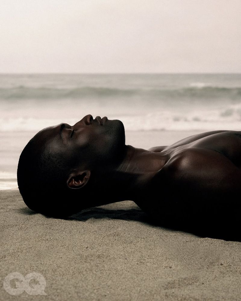 Profile of a shirtless Black man lying on a sandy beach with the ocean in the background for GQ Turkey.