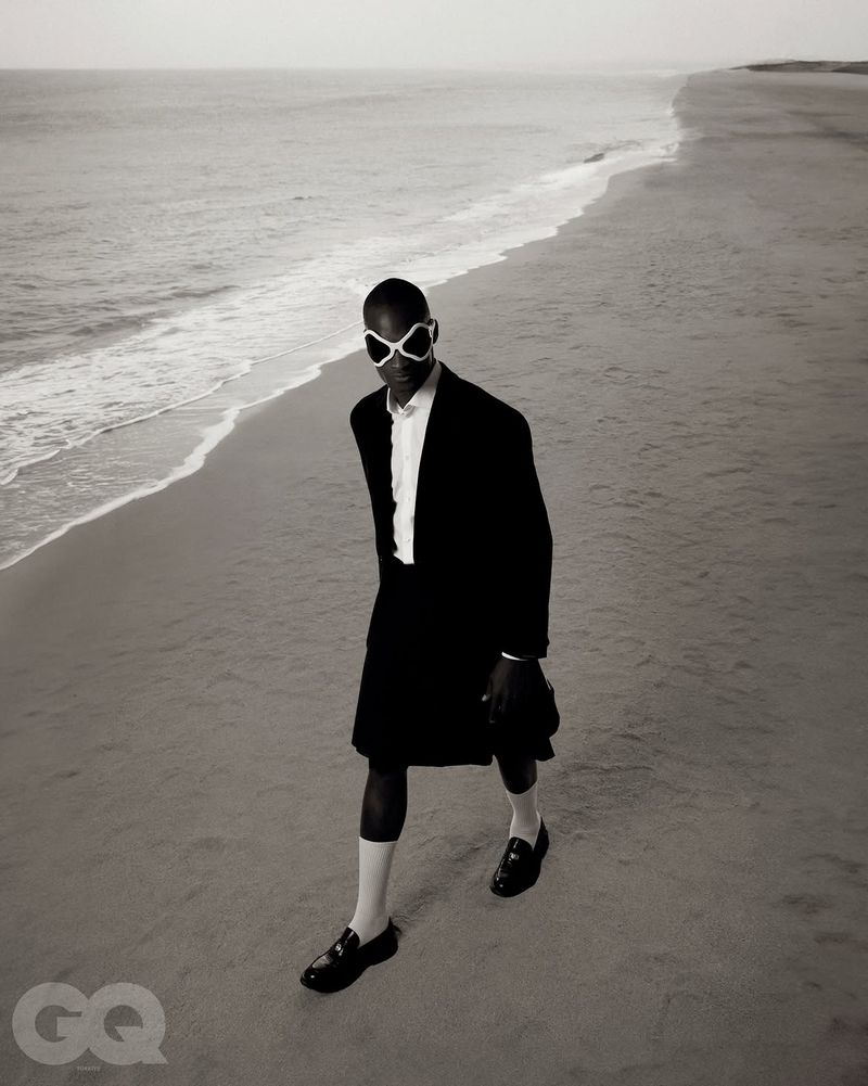 Model wearing dark suit jacket and white sunglasses walking along a sandy beach coastline