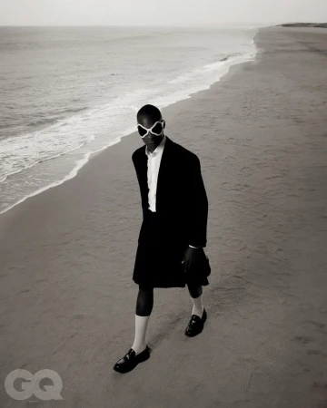 Model wearing dark suit jacket and white sunglasses walking along a sandy beach coastline