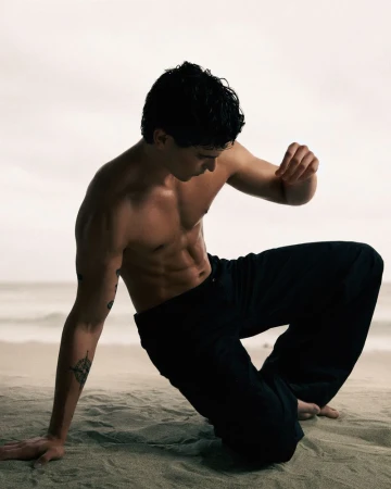 Shirtless male model kneeling on a sandy beach wearing dark trousers under an overcast sky for GQ Turkey