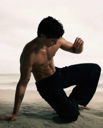 Shirtless male model kneeling on a sandy beach wearing dark trousers under an overcast sky for GQ Turkey