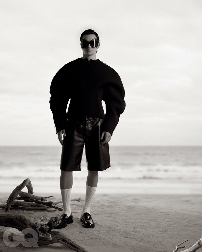Model wearing dark statement sunglasses, black top, leather shorts, and white socks standing on a sandy beach.