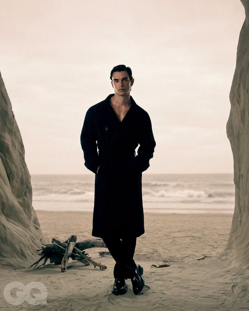 A man wearing a black coat standing on a sandy beach framed by rock formations looking at the camera