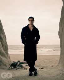 A man wearing a black coat standing on a sandy beach framed by rock formations looking at the camera