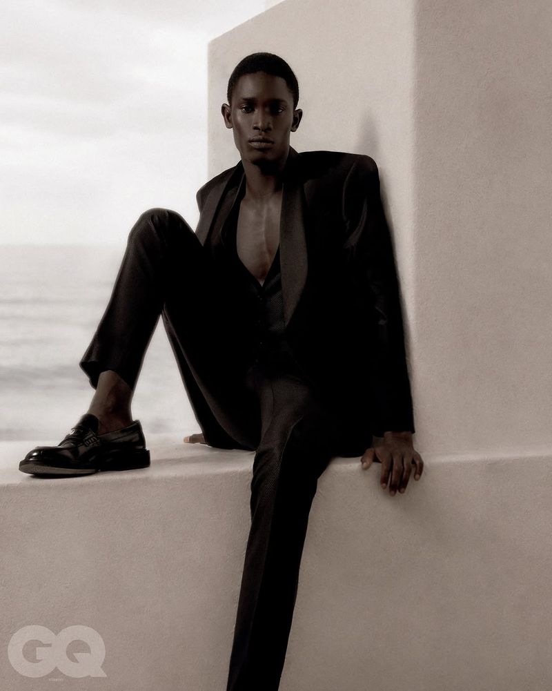 A man in a black suit sitting on a white ledge outdoors against a bright sky for GQ Turkey.