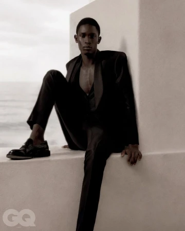 A man in a black suit sitting on a white ledge outdoors against a bright sky for GQ Turkey.