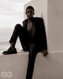 A man in a black suit sitting on a white ledge outdoors against a bright sky for GQ Turkey.
