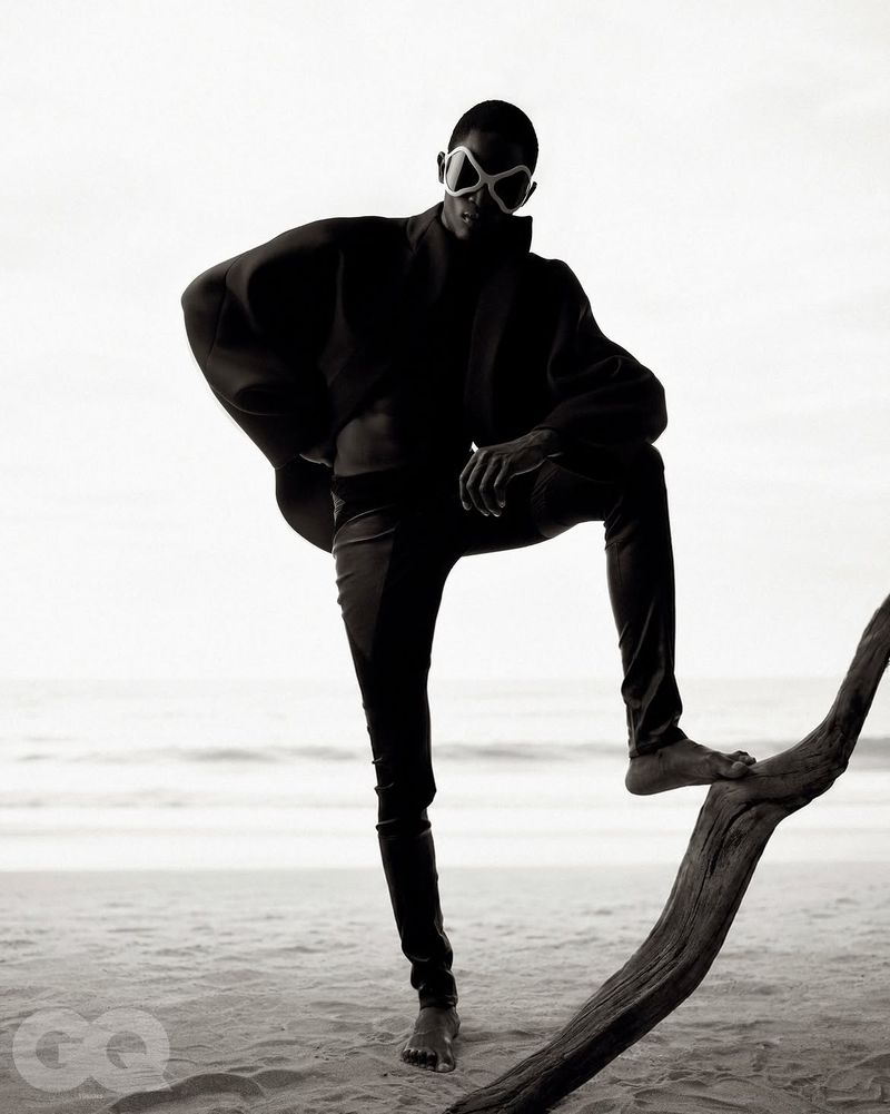Model standing on a sandy beach posing with one foot on a piece of driftwood against an overcast sky.