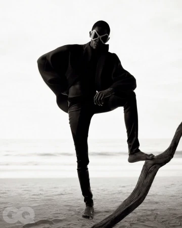 Model standing on a sandy beach posing with one foot on a piece of driftwood against an overcast sky.