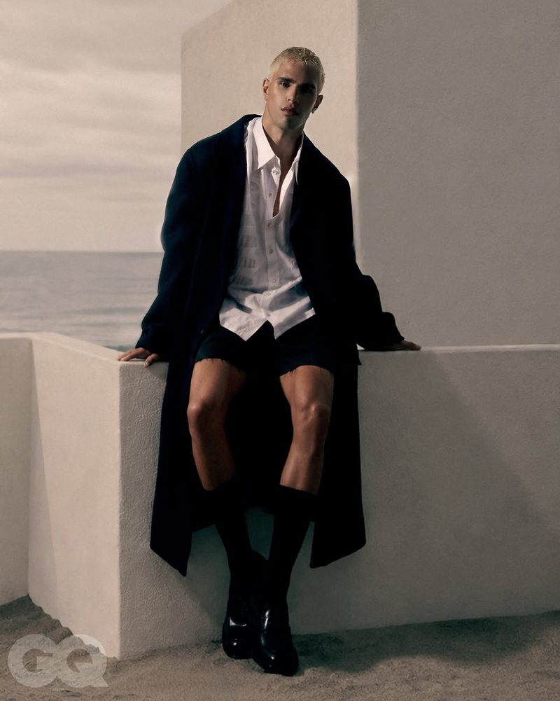 Blonde male model sitting on a white wall by the ocean wearing a dark coat and white shirt for GQ Turkey