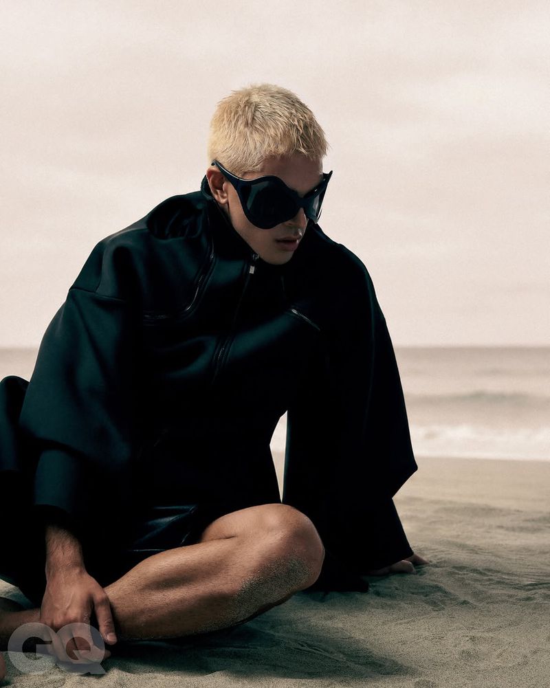 Model with bleached blonde hair wearing black sunglasses and a dark jacket seated on a sandy beach.