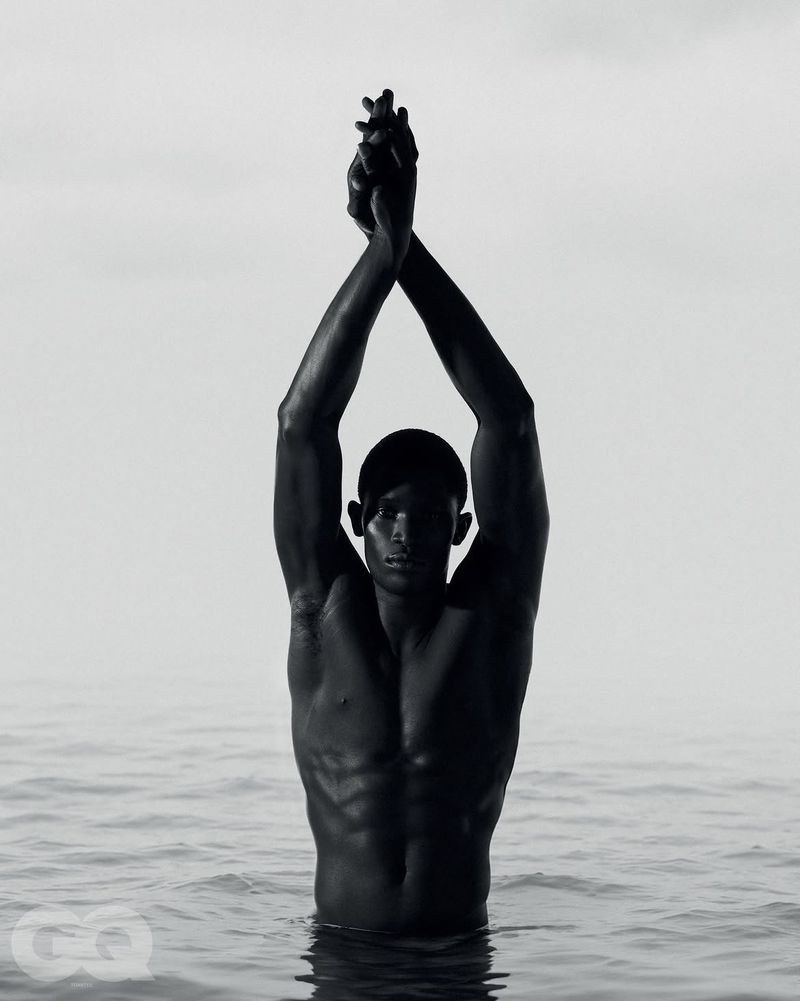 Monochrome portrait of a shirtless man standing waist-deep in water with arms raised and hands clasped overhead.