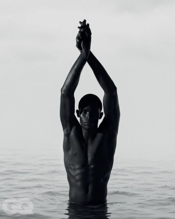 Monochrome portrait of a shirtless man standing waist-deep in water with arms raised and hands clasped overhead.