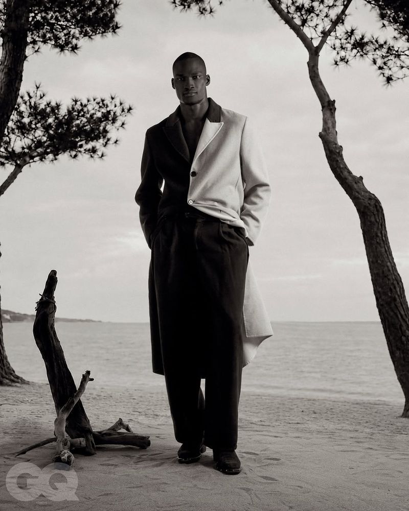 Black model standing on a sandy beach wearing a split black and white coat for GQ Turkey Behind the Sunset editorial.