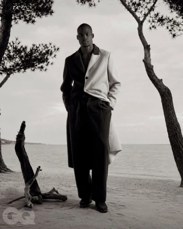 Black model standing on a sandy beach wearing a split black and white coat for GQ Turkey Behind the Sunset editorial.