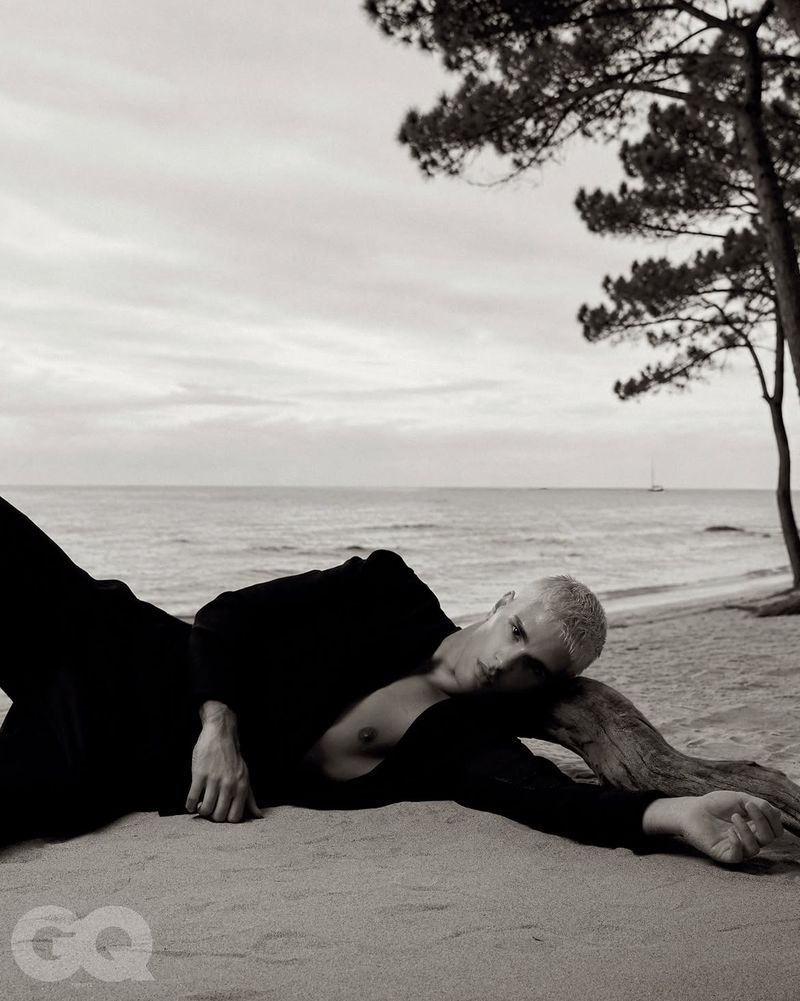 Monochrome fashion editorial image of a male model reclining on a sandy beach resting against a piece of driftwood with the ocean in the background