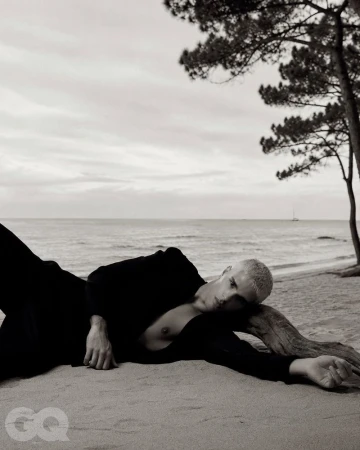 Monochrome fashion editorial image of a male model reclining on a sandy beach resting against a piece of driftwood with the ocean in the background
