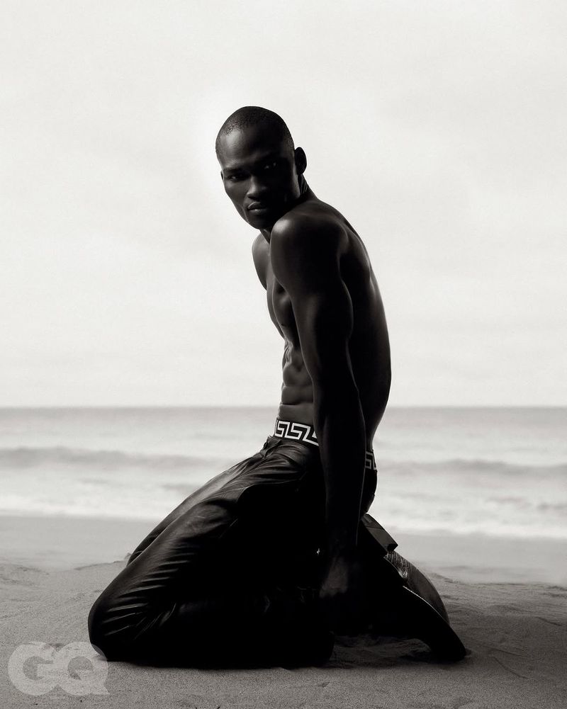 Black male model kneeling on a sandy beach against the ocean, wearing dark patterned trousers.