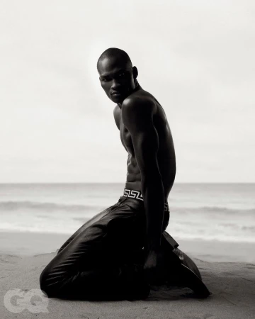 Black male model kneeling on a sandy beach against the ocean, wearing dark patterned trousers.