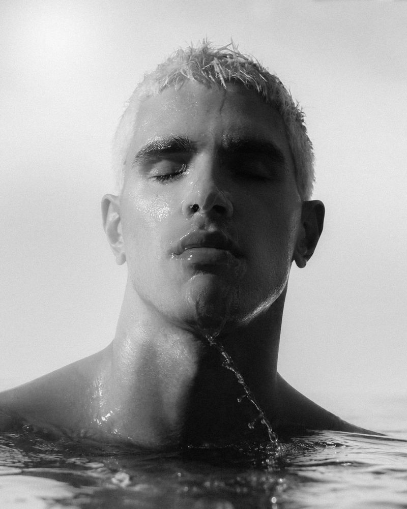 Close-up portrait of a man with light blonde hair standing in water with droplets splashing against his chin and neck.