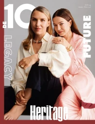 Two women posing together for a fashion magazine cover wearing a white blouse and a pink coat against a grey background.