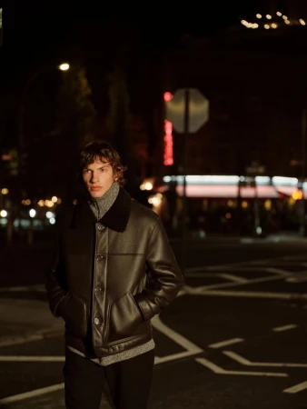 Model wearing a black leather jacket and grey turtleneck standing on a city street at night under ambient lights.