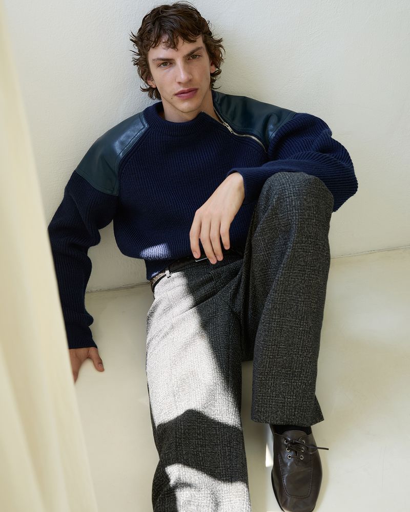 Male model seated against a white wall wearing a navy sweater with dark leather shoulder panels and grey textured trousers