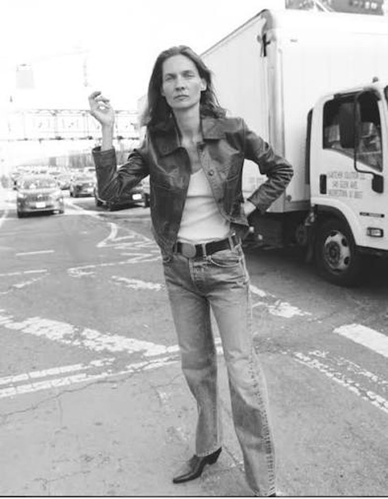 Model wearing a dark leather jacket and denim jeans standing outdoors on a city street beside a parked truck.