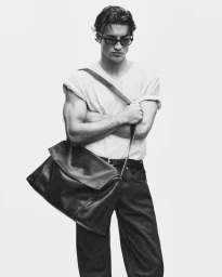 Young man wearing sunglasses and white t-shirt carrying a large leather shoulder bag against a white background.