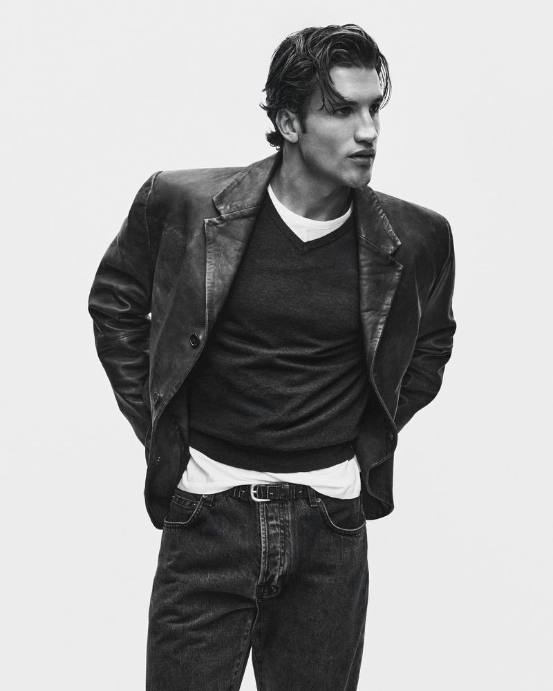 Black and white portrait of a male model wearing a dark leather blazer, V-neck sweater, and dark jeans against a white background.